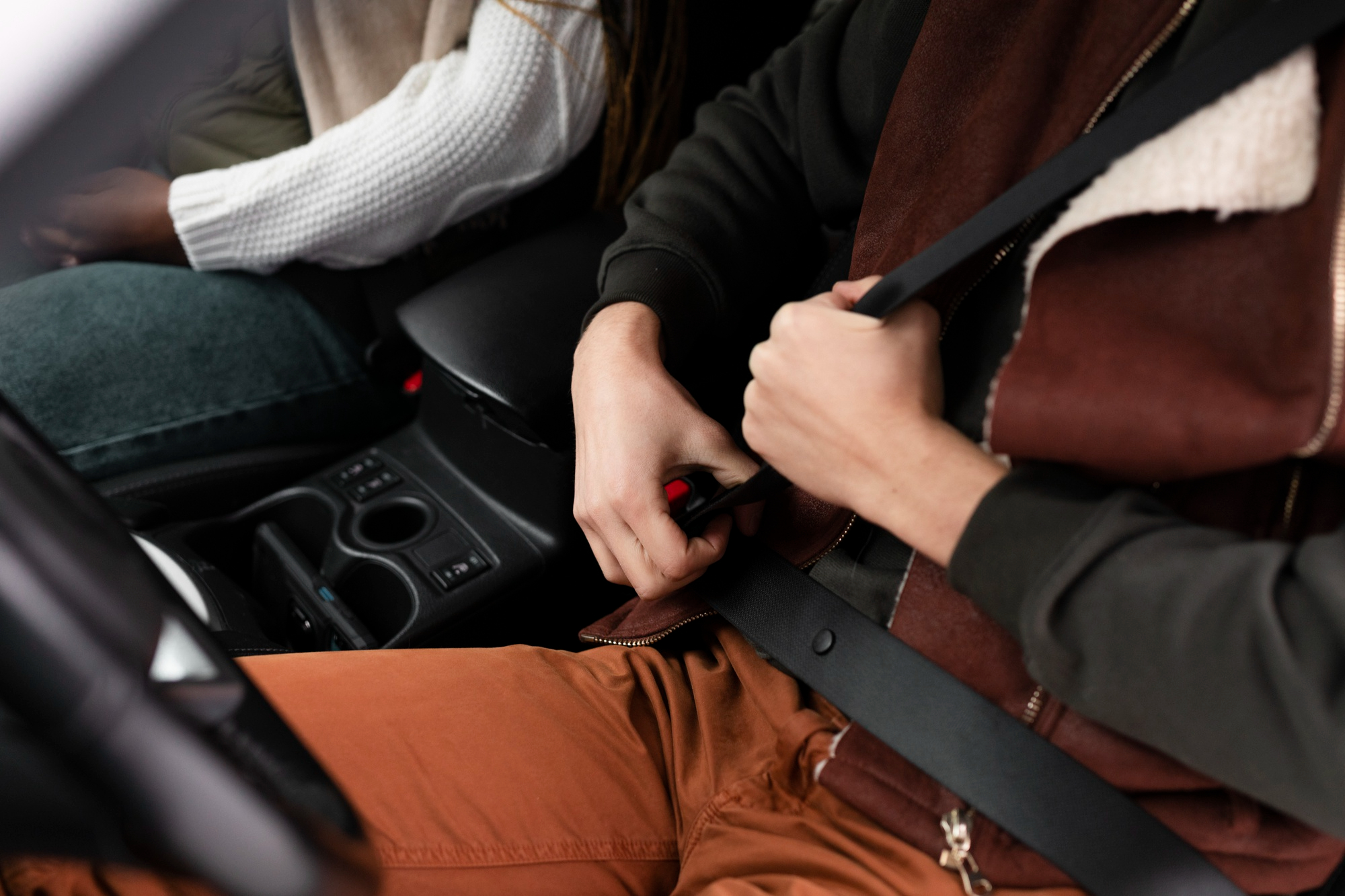 A person in a brown jacket and orange pants fastens their seatbelt in a car, sitting next to another passenger in a white sweater and blue jeans—highlighting the importance of safety to help prevent car accidents NSW. A person in a brown jacket and orange pants fastens their seatbelt in a car, sitting next to another passenger in a white sweater and blue jeans—highlighting the importance of safety to help prevent car accidents NSW.